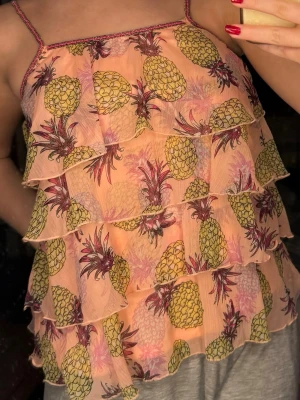 Ananas mönstrad topp - Säljer en söt topp i ananas mönster med tunna axelband och flera volanglager. Toppen har ett färgglatt ananasmönster i gult och lila, och är gjord i ett lätt, transparent syntetmaterial. Perfekt för varma dagar och sommarhäng! Den är i barn storlek EUR 134/140, men jag är 163 centimeter och i 20 års åldern och passar fortfarande perfekt. 