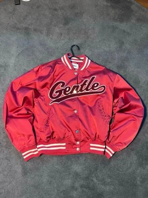 bomberjacka från Weekday - Säljer en röd bomberjacka från Weekday med vit text 'Gentle' på bröstet. Jackan har vita och röda ränder på krage, ärmslut och nederkant, samt knappar framtill. Materialet är blankt och känns syntetiskt, perfekt för dig som gillar sportig och retro stil.