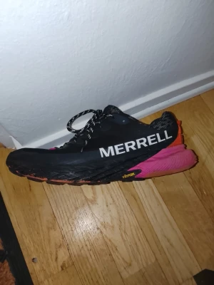 Merrell Agility Peak 5 sneakers - Säljer ett par Merrell Agility Peak 5 sneakers med svart ovandel, stora vita loggor och färgglad sula i rosa och orange. Skorna har snörning med mönstrade svarta och vita skosnören samt Vibram-sula för extra grepp. Perfekta för dig som gillar att vara aktiv utomhus.