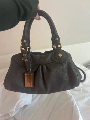 Mörkgrå handväska Marc Jacobs - Säljer en snygg mörkbrun handväska från Marc by Marc Jacobs med guldfärgade metalldetaljer. Den är i jättebra skick, inga defekter alls! 🩷🩷