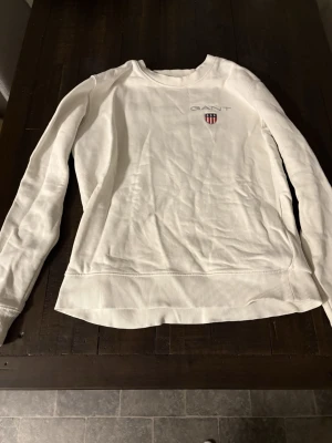 Vit sweatshirt från GANT - Vit långärmad sweatshirt från GANT med rund hals och broderad logga på bröstet. Tröjan har ribbade muddar vid ärmslut och nederkant.