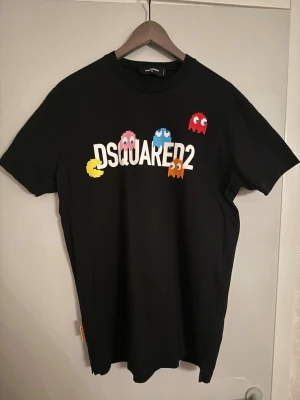Svart DSQUARED2 t-shirt med Pac-Man tryck - Säljer en svart t-shirt från DSQUARED2 med färgglatt Pac-Man tryck framtill. T-shirten har klassisk passform, rund hals och korta ärmar. Materialet är mjuk bomull och trycket har retro gaming-vibe med figurer i gult, rött, blått, rosa och orange.