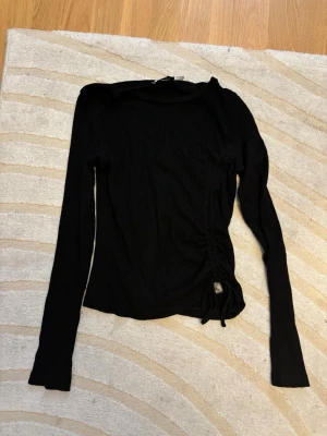 Svart ribbad långärmad topp NA-KD - Säljer en svart ribbad långärmad topp från NA-KD i storlek M. Toppen har snörning på ena sidan som ger en snygg detalj och lite croppad känsla. Materialet är mjukt och stretchigt, perfekt för att styla med jeans eller kjol. Passar mer S