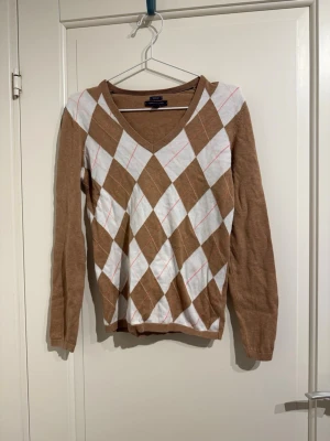 Brun rutig tröja Tommy Hilfiger - Snygg brun och vit långärmad tröja från Tommy Hilfiger med klassiskt rutig argyle-mönster och v-ringad hals. Tillverkad i mjuk pima bomull, med diskret logga på ärmen.