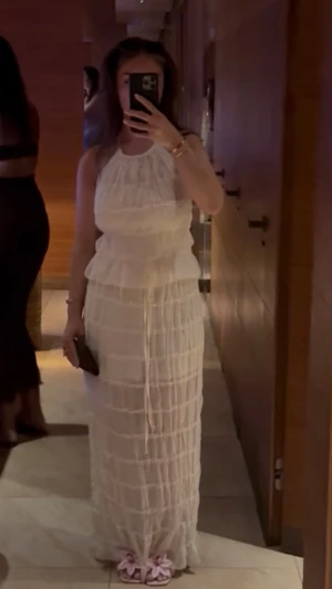 Vit långklänning med volangdetaljer och öppen rygg  - Säljer en superfin vit långklänning med volangdetaljer och halterneck. Klänningen har ett skirt, lätt material och markerad midja med knytband. Perfekt för sommarens alla tillfällen och ger en romantisk vibe.