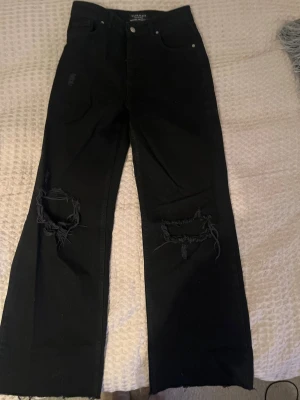 Svarta jeans från Chiquelle - Svarta jeans från Chiquelle med raka ben och råa kanter. Snygga slitningar och hål vid knäna ger en cool vibe. Klassisk femficksmodell och midjehöjd. Materialet är jeans och färgen är helsvart.