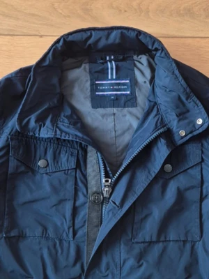 Mörkblå vindjacka från Tommy Hilfiger - Snygg mörkblå vindjacka från Tommy Hilfiger med hög krage, flera fickor med knappstängning och både dragkedja och knappar framtill. Jackan har en clean look och är perfekt för dig som gillar stilrena detaljer och funktion.