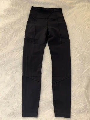 Svarta högmidjade leggings - Snygga svarta leggings med hög midja och markerade sömmar. De har praktiska sidofickor och en tight passform som sitter skönt vid träning eller chill. Materialet känns stretchigt och följsamt. Liten logga framtill i midjan.