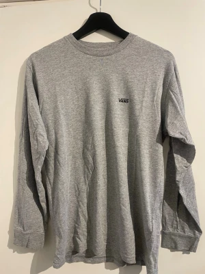Grå långärmad t-shirt från Vans - Säljer en stilren grå långärmad t-shirt från Vans med liten logga på bröstet. Tröjan har rund halsringning och är gjord i mjuk bomull, perfekt för en avslappnad look. Enkel och clean design utan mönster eller extra detaljer.
