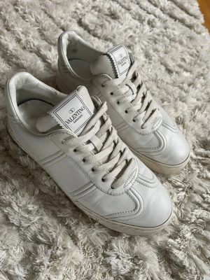 Vita Valentino flycrew sneakers - Snygga vita sneakers från Valentino i skinn med klassisk rund tå, snörning och coola nitar baktill på sulan. Stilren design med diskret logga på plösen och detaljerade sömmar. Skorna är märkta med storlek 36 men passar 37-38. 