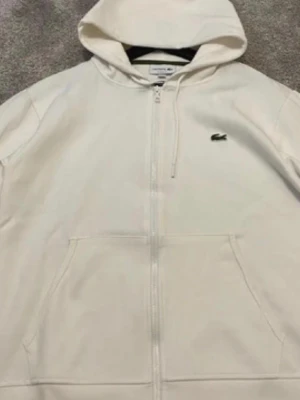 Vit hoodie med dragkedja från Lacoste - Vit hoodie från Lacoste med dragkedja framtill och klassisk krokodillogga på bröstet. Hoodien har kängurufickor och justerbar huva. Tillverkad i mjukt bomullsmaterial för en clean och stilren look.