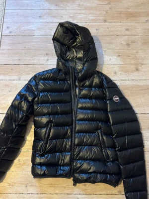 Svart quiltad pufferjacka från Colmar - Snygg svart pufferjacka från Colmar med glansig finish, huva och två dragkedjeförsedda fickor, knappt använd. Jackan har ett quiltat mönster och Colmar-logga på ärmen. 