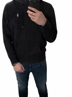 Svart hoodie från Polo Ralph Lauren - Svart hoodie med dragkedja från Polo Ralph Lauren. Klassisk design med vit broderad logga på bröstet, huva med dragsko och kängurufickor framtill. Perfekt för en avslappnad och stilren look.