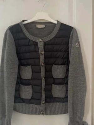 moncler cardigan  - Snygg cardigan jacka från Moncler i grått och svart med quiltad front och fyra stickade fickor. Rund halsringning, ribbade muddar och knappstängning framtill. Perfekt för lager på lager. jackan är i storlek L men är en storlek S enligt mig och sitter litet.