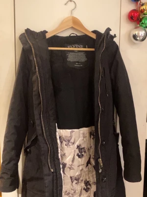 Svart parkas från Elvine med huva - Svart parkajacka från Elvine med huva och dragkedja samt knappar framtill. Jackan har långa ärmar och innerfoder med djurmotiv. Praktiska fickor och justerbar midja med snörning. Perfekt för kyliga dagar.