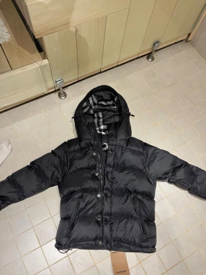Svart dunjacka från Burberry - Snygg svart dunjacka från Burberry med klassiskt rutigt foder i huvan. Jackan har dragkedja och knappar framtill, samt en justerbar huva. Perfekt för kalla dagar och riktigt stilren look.