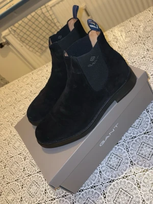 Svarta Chelsea boots från Gant - Stilrena svarta Chelsea boots från Gant i mjuk mocka. Klassisk rund tå, elastiska sidopaneler och dragflikar bak för enkel påtagning. Snygg och tidlös design som passar till det mesta. Levereras i originalkartong.