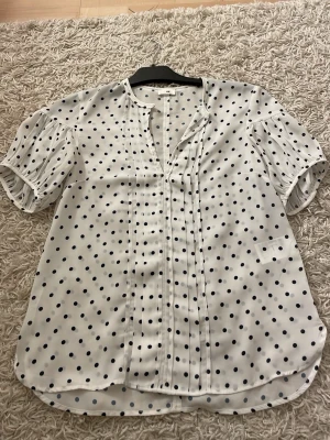 Vit prickig blus från H&M - Söt vit blus från H&M med svarta prickar och korta puffärmar. Blusen har en avslappnad passform, rund halsringning med liten V-öppning och dekorativa veck framtill. Tillverkad i ett lätt och luftigt material som känns skönt mot huden.