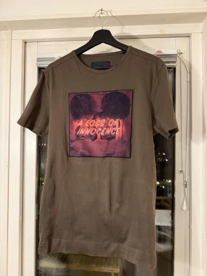 Brun Limitato t-shirt med tryck 'A Loss of Innocence' - Brun Limitato t-shirt med ett stort tryck framtill i rött och svart med texten 'A Loss of Innocence'. T-shirten har rund hals och korta ärmar. Materialet känns som mjuk bomull och passformen är normal. Perfekt för dig som gillar statement-plagg.