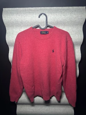 Röd stickad tröja från Polo Ralph Lauren - Snygg röd stickad tröja från Polo Ralph Lauren med rund halsringning och den klassiska blå loggan broderad på bröstet. Tröjan har långa ärmar och ribbade muddar vid ärmslut och nederkant. Perfekt för dig som gillar tidlös och enkel stil.