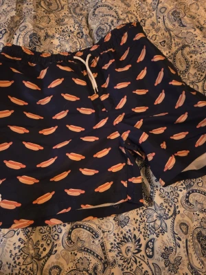 Mörkblå badbyxor med korvtryck - Säljer ett par mörkblå badbyxor med elastisk midja och vit dragsko. Badbyxorna har ett roligt mönster med korvar i bröd över hela tyget. Perfekta för stranden eller poolen om du vill sticka ut! Materialet känns snabbtorkande och lätt.