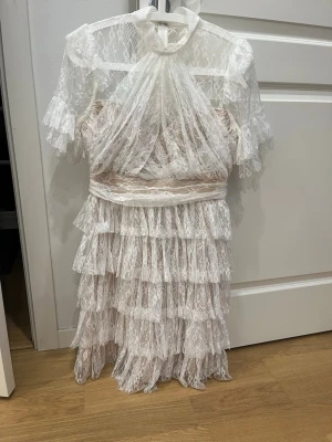 Vit spetsklänning med volanger från By Malina - Superfin kort vit klänning i spets med volanger och transparenta detaljer. Hög hals, korta ärmar och omlottliknande överdel. Klänningen har flera lager spets och en romantisk vibe, perfekt för dig som vill sticka ut. Perfekt till studenten! Använd endast 1 gång 