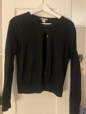Svart kofta från H&M BASIC - En klassisk svart kofta från H&M BASIC med rund halsringning och knäppning framtill. Långärmad modell i mjukt och stretchigt material, perfekt att slänga över en t-shirt eller linne. Enkel och stilren design som funkar till det mesta.