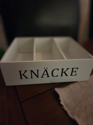 Vit trälåda med texten KNÄCKE - Vit rektangulär trälåda med tre fack och svart text 'KNÄCKE' på framsidan. Perfekt för att förvara knäckebröd eller andra småsaker på köksbordet. Lådan har en stilren och enkel design.