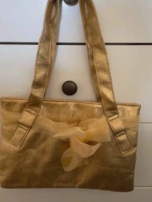 Guldig handväska med rosett - Stilren guldig handväska med dubbla axelband och en dekorativ rosett framtill. Väskan har en rektangulär form och är perfekt för dig som vill sticka ut med en glansig accessoar. Passar till många olika outfits och ger en lyxig känsla. Knapplås. Ingen dragkedja.