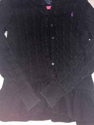 Svart kabelstickad kofta Ralph Lauren - Kabelstickad svart kofta från Ralph Lauren med lila broderad logga på bröstet. Koftan har långa ärmar, ribbade muddar och knäppning framtill. Klassisk och tidlös design som passar till mycket. Originalpris 2300. Aldrig andvänd