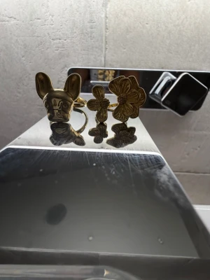 Två guldiga statementringar - Säljer två unika ringar i guldfärgad metall, en med ett detaljerat hundhuvud och en med två blommor i olika storlekar. Perfekta för dig som vill sticka ut och addera en lekfull touch till din stil. Jag är öppen för prisförslag. Jag säljer dessa ringar eftersom att dom inte passar mig. Dessa ringar är helt nya och i nytt skick. Jag säljer dessa för 100kr för båda och jag köpte dom för 330kr🤍