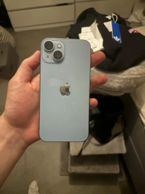 Ljusblå iPhone 14 med dubbla kameror - Säljer en ljusblå iPhone 14 från Apple. Telefonen har en stilren design med platt baksida i glas, aluminiumram och två stora kameralinser i hörnet. Perfekt för dig som vill ha en modern och snygg mobil med grym kamera och klassisk Apple-känsla.