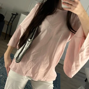 Ljusrosa topp med vida ärmar och rosett - Söt ljusrosa topp med trekvarts vida ärmar och stor rosett i ryggen. Toppen har en lös passform och är tillverkad i ett mjukt, tunt tyg som ger ett flowy intryck. Perfekt för dig som gillar feminina detaljer och en avslappnad men trendig stil.