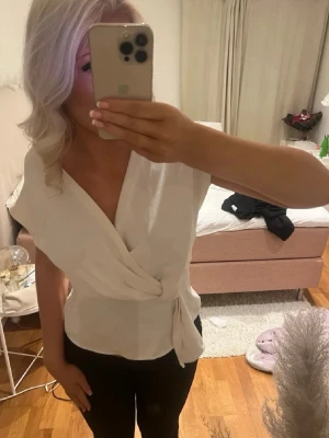 Vit omlottopp med knyt i midjan - Snygg vit omlottopp med knyt i midjan och v-ringad hals. Toppen har korta ärmar och en stilren, feminin look som passar perfekt till både jeans och kjol. Enkel att matcha och superfräsch till sommaren. Sömnen har släppt lite på ena armen men inget man tänker på