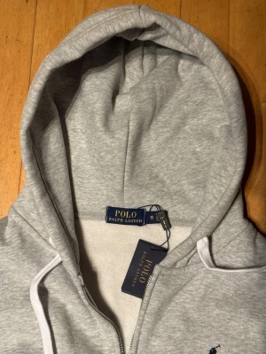 Grå Zip up från Polo Ralph Lauren - Snygg ljusgrå hoodie från Polo Ralph Lauren med dragkedja och vit dragsko i huvan. Klassisk broderad logga i marinblått på bröstet. Tillverkad i mjukt bomullsmaterial som känns riktigt skönt mot huden. Perfekt för en chill och stilren look.