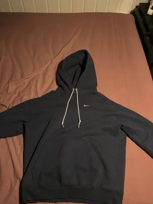 Mörkblå hoodie från Nike - Snygg mörkblå hoodie från Nike med klassisk huva och vita dragsnören. Liten Nike-logga broderad på bröstet och stor känguruficka framtill. Perfekt för en avslappnad och sportig stil. Tillverkad i mjukt material för extra komfort. Oversize så den passar till de som har storlek L
