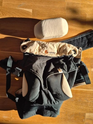 Svart bärsele från Ergobaby - Praktisk svart bärsele från Ergobaby med beige detaljer och vadderade axelband. Selen har justerbara remmar och medföljer ett mjukt nackstöd i beige tyg. Perfekt för att bära små barn på ett bekvämt sätt. Funkar från 0 till 36 mån.
