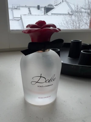 Dolce & Gabbana parfym (tom flaska) - Säljer en tom flaska Dolce & Gabbana Rosa Excelsa parfym i en glasflaska med elegant svart rosett runt halsen. Flaskan har ett unikt lock i form av en röd ros som ger en lyxig känsla. Den kan användas som dekoration eller samlarobjekt #dolce&gabbana #parfym