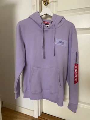 Hoodie Alpha Industries - Snygg ljuslila hoodie från Alpha Industries med stor vit logga på ryggen och mindre sopatch framtill. Klassisk känguruficka, ribbade muddar och huva med dragsko. Ärmen har en ficka med dragkedja och röd tagg. Perfekt för en avslappnad streetwear-look.