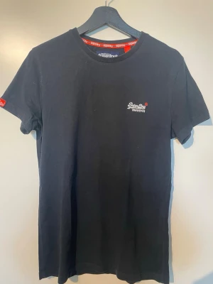 Svart t-shirt från Superdry - Klassisk svart t-shirt från Superdry med liten logga broderad på bröstet och röd detalj på ärmen. T-shirten har rund halsringning och är tillverkad i mjuk bomull. Perfekt basplagg med normal passform och korta ärmar.