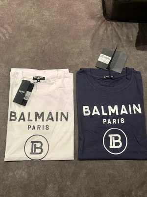 2X  vit och blå Balmain Paris t-shirt med logga - Säljer två snygga Balmain Paris t-shirts i klassisk modell. Ena är vit och den andra är mörkblå, båda med stor Balmain-logga och tryck framtill. T-shirtsen har rund hals och korta ärmar. Perfekt för dig som gillar stilrena och trendiga plagg.