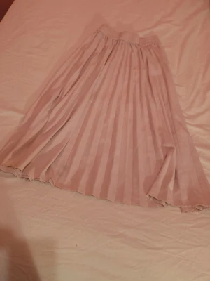 Ljusrosa plisserad långkjol - Säljer en ljusrosa långkjol med plisserad design och elastisk midja. Kjolen har ett mjukt, flytande tyg som ger en snygg siluett och passar perfekt till olika stilar. Superfin att matcha med både sneakers och boots.