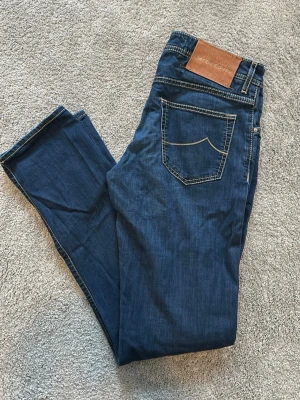 Blå jeans från Jacob Cohen  - Snygga blå jeans från Jacob Cohen, modell NICK, med klassisk femficksmodell och kontrastsömmar. Jeansen har en rak slim fit passform och patch i brunt läder bak i midjan. Tillverkade i mjukt denimtyg som ger en skön känsla och stilren look. Mycket bra skick på jeansen, använda fåtal gånger. Storlek 31. Midjemått = 40 cm. Ytterbenslängd = 104 cm.