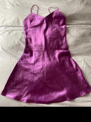 Lila satin klänning med smala band - Säljer en lila kortklänning i satin med smala axelband och lätt utsvängd kjol. Klänningen har en enkel, elegant design med slät yta och figurnära passform upptill. Perfekt för dig som vill ha en stilren och trendig look.