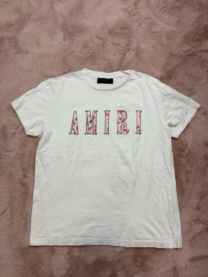 AMIRI t-shirt - Säljer en vit t-shirt från AMIRI med korta ärmar och ett stort blommigt AMIRI-tryck i rött och rosa på bröstet. T-shirten har en klassisk passform och är gjord i mjuk bomull, perfekt för en avslappnad och trendig look.