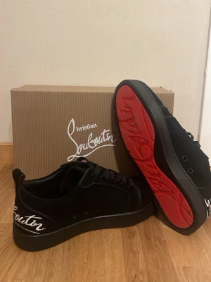 Svarta sneakers från Christian Louboutin - Snygga svarta sneakers från Christian Louboutin med klassisk röd sula och vit broderad logga på sidan. Skorna har snörning och en stilren design som sticker ut med den ikoniska röda undersidan. Pris kan såklart diskuteras.
