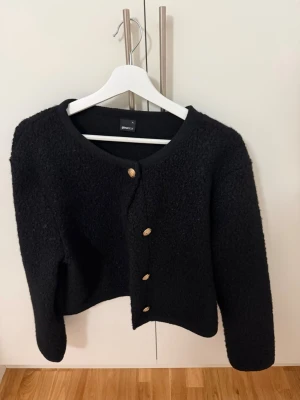 Svart jack - Säljer en svart bouclékofta från Gina Tricot med rund halsringning och fyra guldfärgade knappar framtill. Perfekt att styla till jeans eller kjol för en trendig look. Använd en gång💞