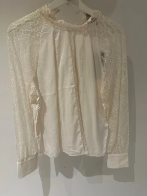 Vit spetsblus med lång ärm Vila Rouge storlek 40 - Säljer en elegant vit/beige blus med transparenta spetsärmar och hög hals. Blusen har ett romantiskt mönster i spetsen och släta manschetter. Perfekt för dig som gillar en feminin och trendig stil. Materialet är en mix av spets och tyg. Knäppning bak i nacken.