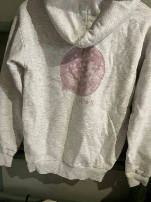 Ljusgrå hoodie från Gina Tricot - Ljusgrå hoodie från Gina Tricot med en stor rosa discokula och texten ICXN på ryggen. Hoodien har ribbade muddar vid ärmslut och nederkant samt en klassisk huva. Tillverkad i mjukt sweatshirtmaterial som är skönt att ha på sig.