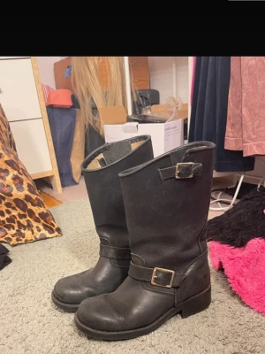 Svarta skinnstövlar med spännen. Johnny Bulls - Svarta stövlar i skinn från Henry Shoes med två snygga spännen och robust sula. Klassisk rund tå och låg klack, perfekt för dig som gillar en tuffare stil. Stövlarna har en enkel och stilren design som passar till många outfits.